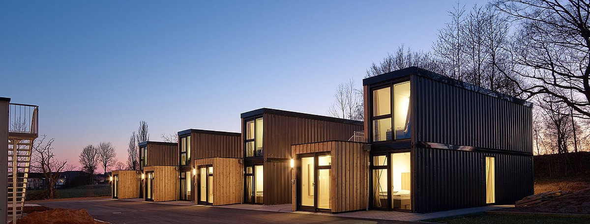 Moderne Wohncontainer aus Holz und Metall mit leuchtenden Fenstern bei Abenddämmerung, umgeben von Bäumen und beleuchtetem Vorplatz.