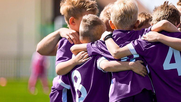 Kinder mit violetten Trikots einer Fußballmannschaft, die Arm in Arm im Kreis auf einem Fußballplatz stehen.
