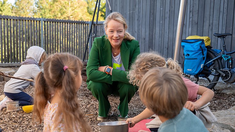 Umweltministerin Thekla Walker sitzt in der Hocke und spricht mit Kindern im Garten.