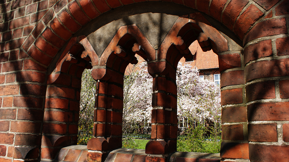In einer Backsteinmauer öffnet ein gemauerter Bogen den Blick auf blühende Obstbäume. Im Bogen befinden sich zwei Säulen, die den Bogen tragen.