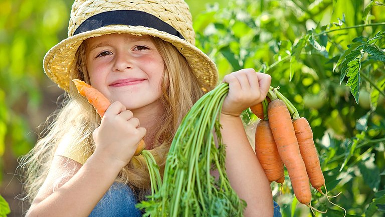 Ein Mädchen mit einem Strohhut auf dem Kopf hält in einer Hand ein Bund Karotten in der anderen eine einzelne.