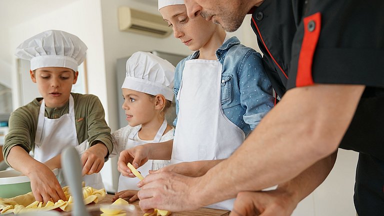Drei Kinder im Grundschulalter, die mit einem erwachsenen Koch einen Teig in einer Springform mit Äpfeln belegen.
