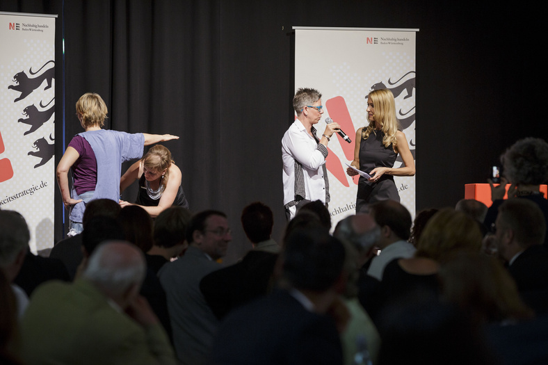 Nachhaltigkeitstage 2014 Auf der Bühne spricht eine Frau zur Moderatorin. Neben ihr stehen zwei Frauen. Es wird ein Kleidungsstück anprobiert/angepasst.