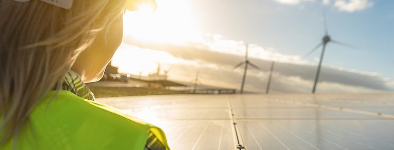 Im Vordergrund sieht man die Rückseite des Oberkörpers eine Frau. Hinten mittig im Bild scheint die Sonne und es werden Windräder gezeigt. Die Frau schaut diesen entgegen.