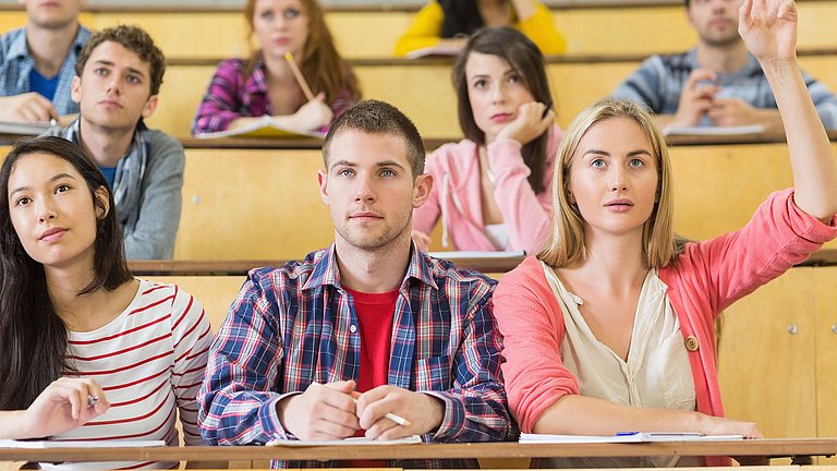 Neun junge Erwachsene in einem Hörsaal, die interessiert nach vorne Blicken. Eine Studentin hebt den Arm nach oben, um etwas zu sagen.