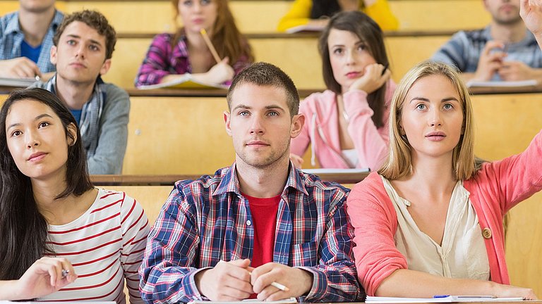 Neun junge Erwachsene in einem Hörsaal, die interessiert nach vorne Blicken. Eine Studentin hebt den Arm nach oben, um etwas zu sagen.