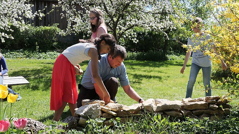 Ein Mann und eine Frau kümmern sich um eine Trockensteinmauer. Im Hintergrund gehen Kinder durch eine große blühenden Freifläche.