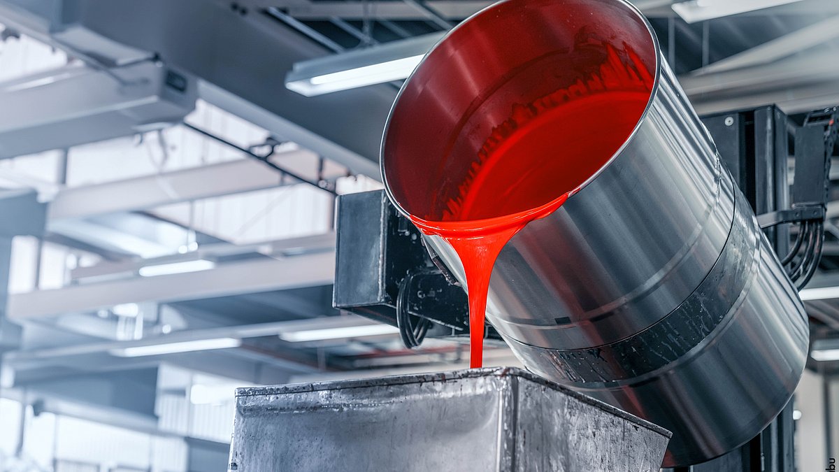 In einer hellen Produktionshalle fließt aus einem großen Metallfass rote Farbe in den Container einer Abfüllanlage.