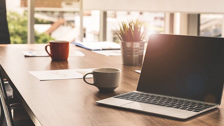 Auf einem Holztisch mit Stühlen drumherum steht ein Laptop, daneben Tassen und ein Blatt Papier.