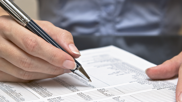 Hand hält einen Stift in der Hand, darunter liegt ein beschriebenes Blatt Papier.