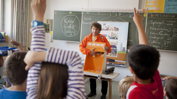 Energie-Detektiv EDe steht mit seiner orangefarbenen Jacke von der Tafel in einem Klassenzimmer und erklärt den Schulkindern einen Lampenkasten, mit dem die Kinder verschiedene Beleuchtungsmittel auf Stromverbrauch und Lichtausbeute untersuchen können. Im Vordergrund sient man ein paar Kinder, die sich eifrig zu Wort melden wollen.
