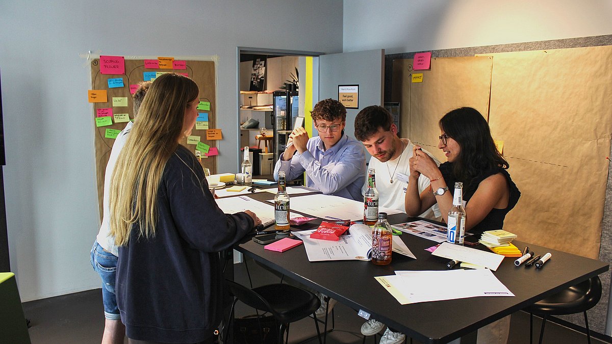 Fünf Personen stehen zentral im Bild um einen Tisch. Es liegen viele Zettel und Post-Its auf dem Tisch und es wird gemeinsam etwas bearbeitet.