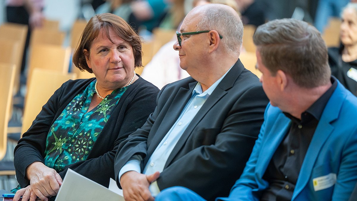 Farbige Fotografie: Drei Personen sitzen in einer Stuhlreihe und sprechen miteinander. Die Frau links hält Dokumente in der Hand.