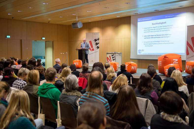 Ein Saal mit Stuhlreihen, eime Bühne mit orangefarbenen N-Würfeln, die Reihen sind voll besetzt. Ein Vortragender steht am Pult. 