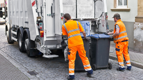 Zwei Männer von der Müllabfuhr in orangenfarbener Kleidung stellen Mülleimer zur Entleerung an einem Müllfahrzeug bereit.