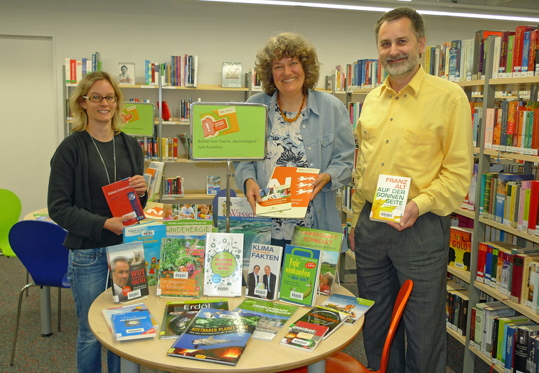 Büchertisch in der Bibliothek. Drei Personen stehen daneben und dahinter und halten Bücher in der Hand.