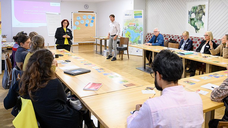 Mehrere Personen sitzen an U-förmig aufgestellten Tischen in einem Workshop, zwei Personen stehen vorne und richten den Blick auf die Menge.