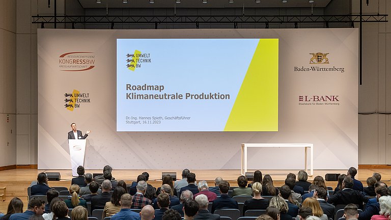 Foto eines Vortrags der Umwelttechnik BW zum Thema klimaneutrale Produktion. Der Geschäftsführer Dr.-Ing. Hannes Spieth steht auf einer Bühne in einem Konferenzraum und spricht zum Publikum.