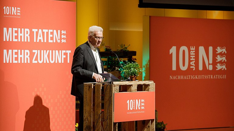 Ministerpräsident Winfried Kretschmann hält an einem Rednerpult eine Rede. Im Hintergrund sind auf orangefarbenen Grund die Schriftzüge "Mehr Taten mehr Zukunft" sowie 10 Jahre Nachhaltigkeitsstrategie zu lesen.
