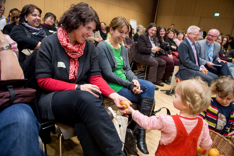 Ein kleines Kind teilt zusammen mit einem anderen Kind Äpfel ans Publikum. Eine Frau freut im Publikum freut sich über den Apfel. Die Frau neben ihr lacht auch.