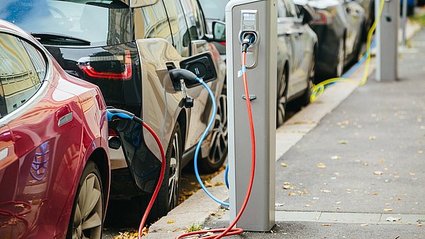 Das Bild zeigt mehrere Elektro-Autos am Straßenrand, die an einer Elektroladesäule angeschlossen sind.