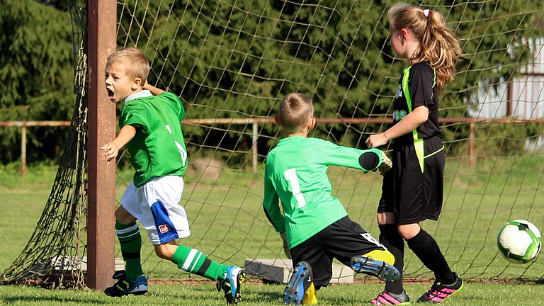 Drei Kinder eines Fußballvereins spielen Fußball. Ein Junge jubelt vor dem Tor. 