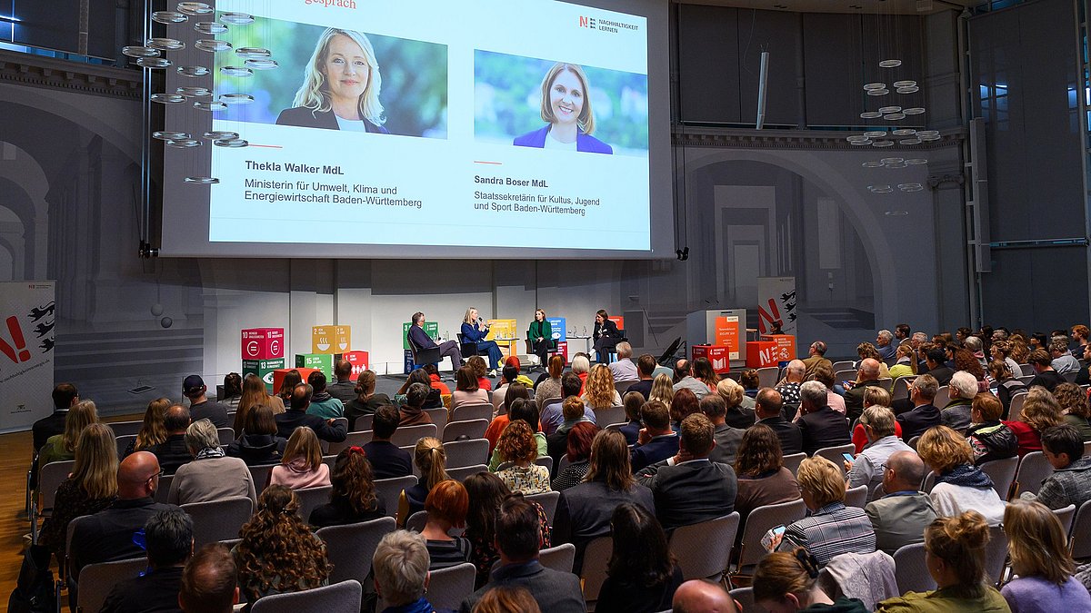Auf der Bühne findet eine Podiumsdiskussion statt zwischen der Umweltministerin Thekla Walker und der Staatssekretärin für Kultur, Jugend und Sport in Baden-Württemberg Sandra Boser. Über Ihnen ist eine große Leinwand auf der eine Präsentation zu sehen ist. Im Vordergrund sind die mit den Zuhörenden gefüllten Stuhlreihen.
