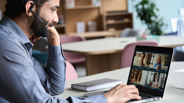 Ein Mann sitzt vor einem mit einem Headset im Ohr Laptop und nimmt an einem Online-Meeting teil. Auf dem Bildschirm sind die Bilder von vier weiteren Personen zu sehen, die am Meeting teilnehmen. 
