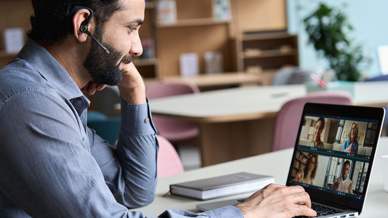 Ein Mann sitzt vor einem mit einem Headset im Ohr Laptop und nimmt an einem Online-Meeting teil. Auf dem Bildschirm sind die Bilder von vier weiteren Personen zu sehen, die am Meeting teilnehmen.