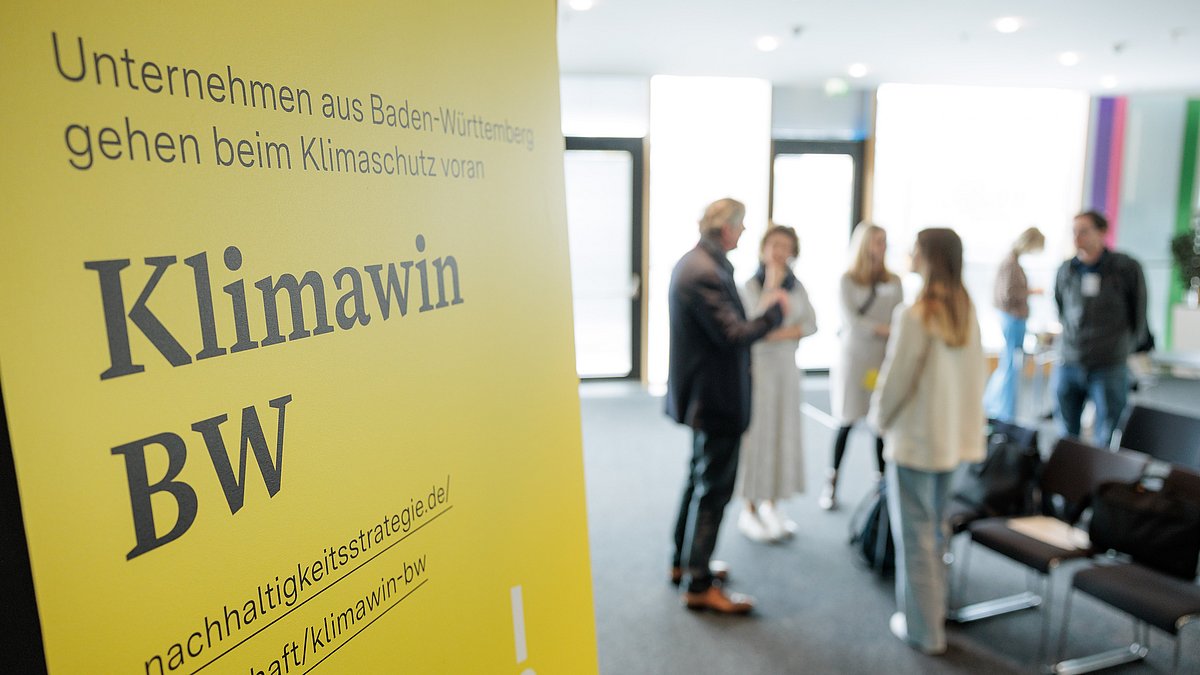 Im Vordergrund ein Aufsteller der Klimawin BW, im Hintegrund eine Gruppe von Teilnehmenden im Austausch.