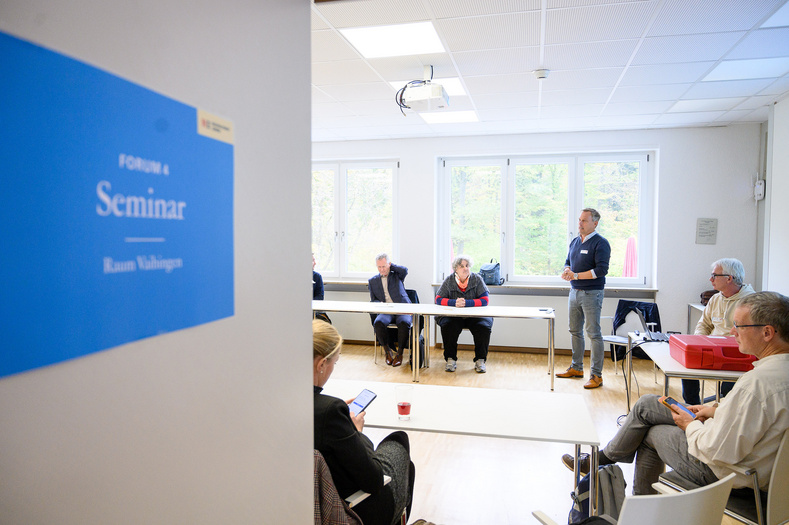 Links im Bild ist ein Ausschnitt einer offenen Tür zu sehen, an der ein hellblaues Schild mit der Aufschrift "Seminar" hängt. Im Hintergrund sieht man einen Raum, in dem mehrere Personen sitzen und einem Redner zuhören.