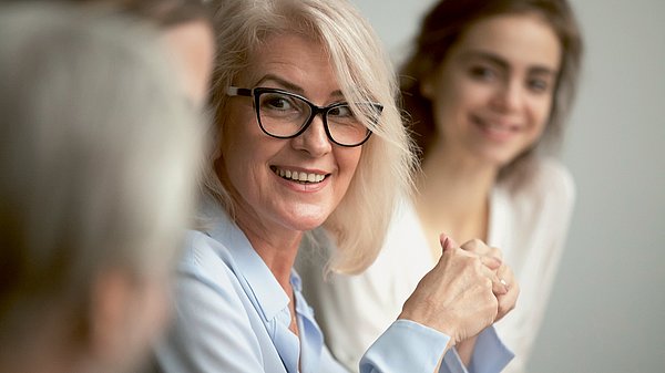 Dargestellt ist eine Besprechung, im Vordergrund eine etwas ältere Frau, die jemandem zuhört. Im Hintergrund eine jüngere Frau.