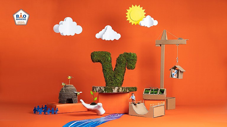 Vor einem roten Hintergrund ist ein V aus grünen Blättern, ein Kran, Wolken, eine Sonne, ein Bienenstock, ein Fluss, Menschen, eine Hand mit einer Pflanze sowie eine Skaterrampe mit einem Playmobil-Männchen aus Papier und anderen Materialien aufgebaut.