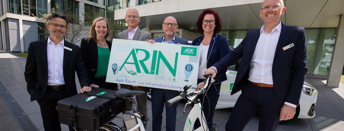 Die AOK Baden-Württemberg stellt Verkehrsminister Winfried Hermann ihr Pilot-Projekt ARIN vor. Die Personen-Gruppe hält ein Schild hoch, davor steh ein AOK-Pedelec.