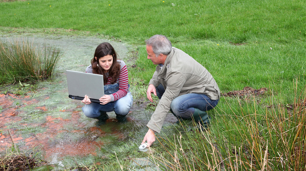 Eine Jugendliche und ein älterer Mann entnehmen auf einer überschwemmten Wiese eine Wasserprobe. Die junge Frau hat einen Laptop in den Händen.