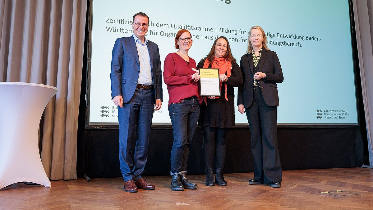 Umweltministerin Thekla Walker MdL und Kultusstaatssekretär Volker Schebesta MdL stehen zusammen mit zwei Frauen auf einer Bühne. Sie lächeln alle in die Kamera, die zwei Frauen halten ein eingerahmtes BNE-Zertifikat.