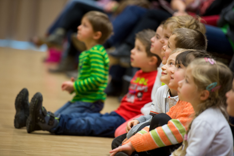 Acht Kinder im Kindergartenalter sitzen auf dem Boden vor dem Publikum und schauen Richtung Bühne.