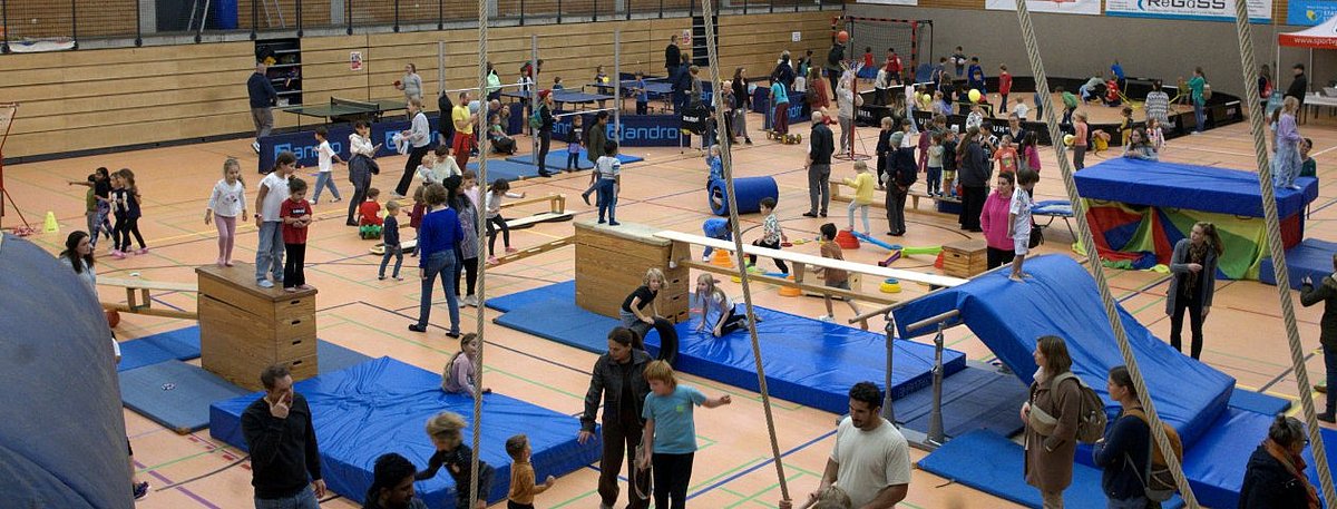 Turnhalle in der viele Menschen sind und verschiedene Stationen eines Parkours mit blauen Matten und Sprungelementen machen