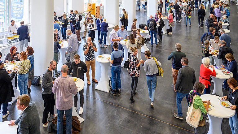 Ein Raum mit vielen runden Stehtischen ist gefüllt von Menschen, die sich gerade am Catering bedienen und ihr Mittagessen genießen.