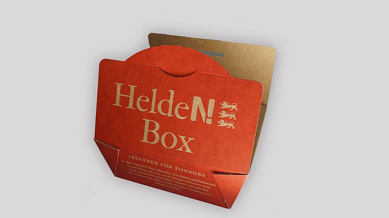 Dargestellt wird eine geöffnete und eine geschlossene Helden-Box aus lebensmittelechtem Karton, in der übrig gebliebenes Essen eingepackt und mit nach Hause genommen werden kann.