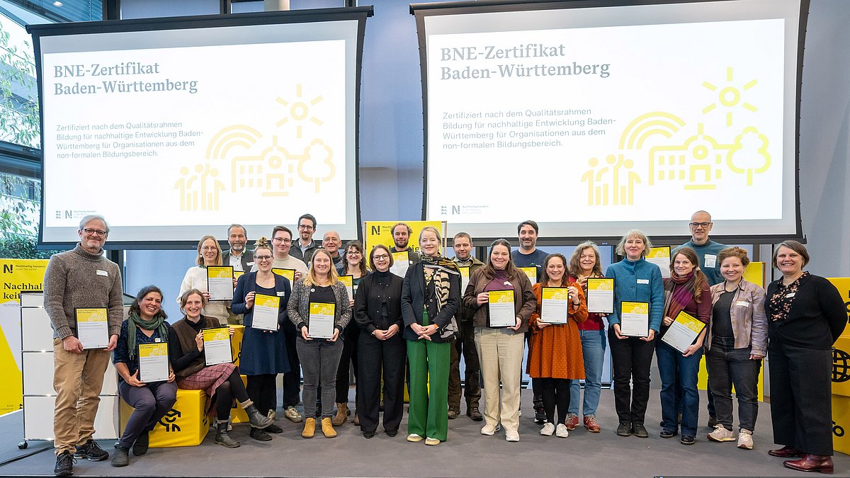 Alle Personen, die eine BNE-Zertifizierung erhalten haben, stehen gemeinsam mit ihrer Urkunde in den Händen mit Umweltministerin Thekla Walker und Kultusstaatssekretärin Sandra Boser auf der Bühne.
