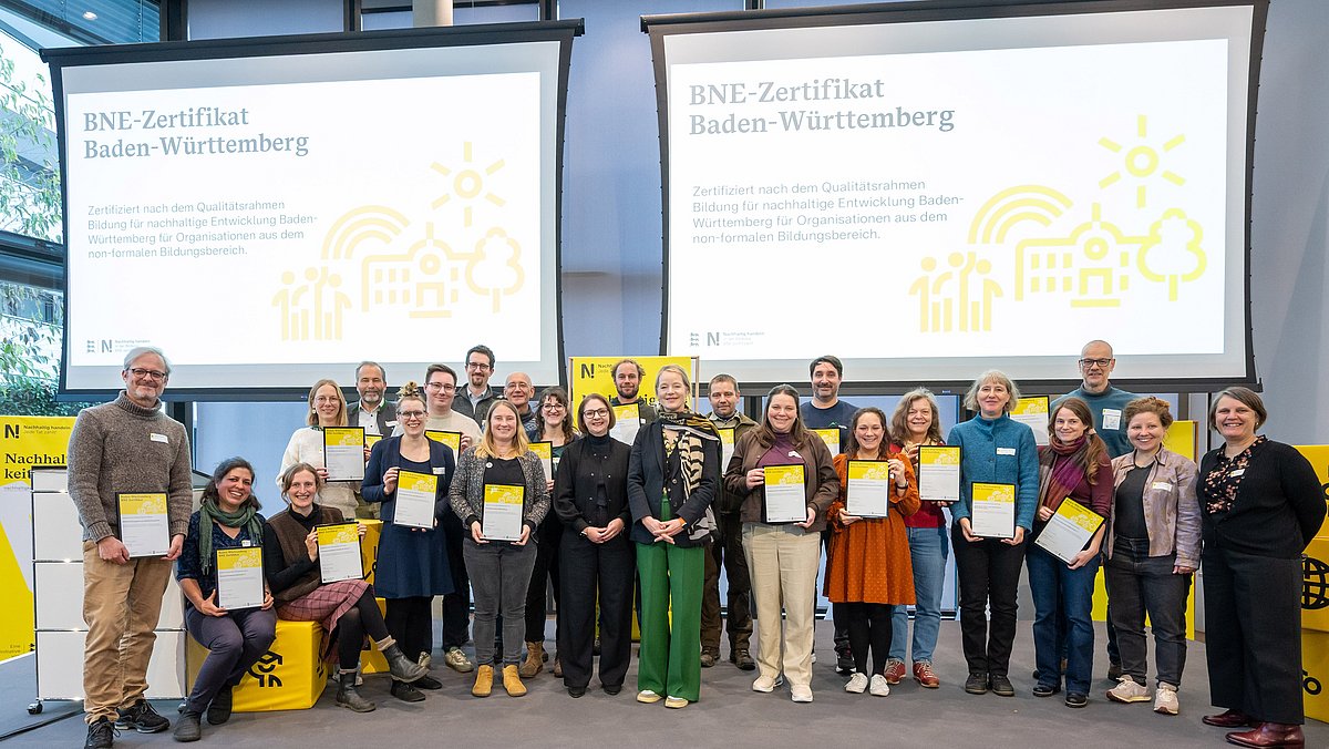 Alle Personen, die eine BNE-Zertifizierung erhalten haben, stehen gemeinsam mit ihrer Urkunde in den Händen mit Umweltministerin Thekla Walker und Kultusstaatssekretärin Sandra Boser auf der Bühne.