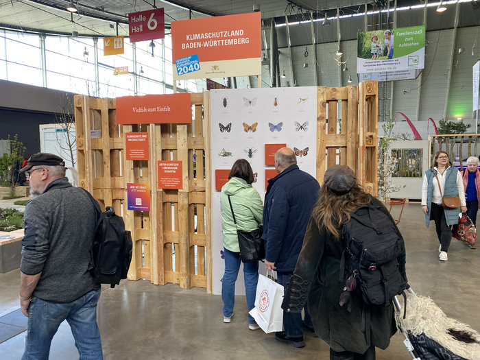 Mehrere Personen betrachten einen Informationsstand aus Paletten, auf dem ein Schild mit Schmetterlingen abgebildet ist.