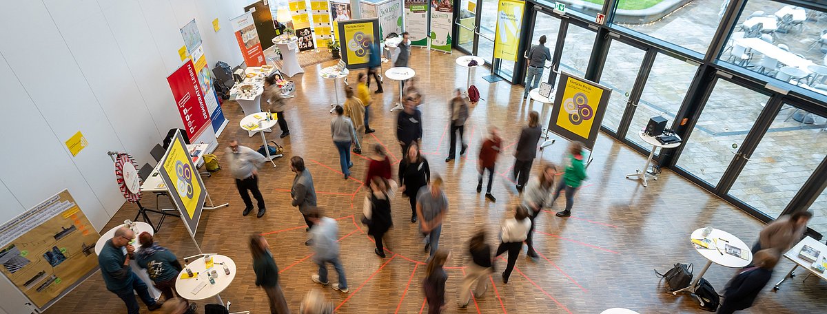 Luftaufnahme eines Raumes mit verschiedenen Ständen der Akteurinnen und Akteure des Markt der Möglichkeiten. Es herrscht reges Treiben.