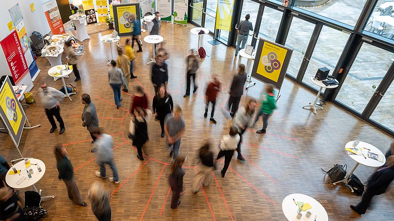 Luftaufnahme eines Raumes mit verschiedenen Ständen der Akteurinnen und Akteure des Markt der Möglichkeiten. Es herrscht reges Treiben.