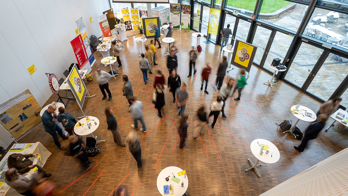 Luftaufnahme eines Raumes mit verschiedenen Ständen der Akteurinnen und Akteure des Markt der Möglichkeiten. Es herrscht reges Treiben.