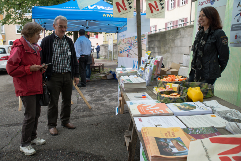 Ein Stand mit Broschüren, dahinter einer Frau, davor zwei Personen.