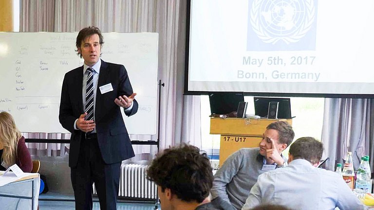 Mehrere junge Erwachsene an zwei Tischen, die dem vortragenden Mann in der Mitte des Raums zuhören