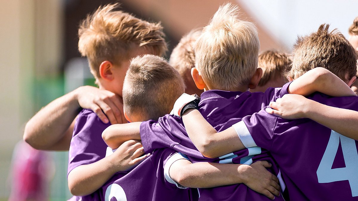 Kinder mit violetten Trikots einer Fußballmannschaft, die Arm in Arm im Kreis auf einem Fußballplatz stehen.