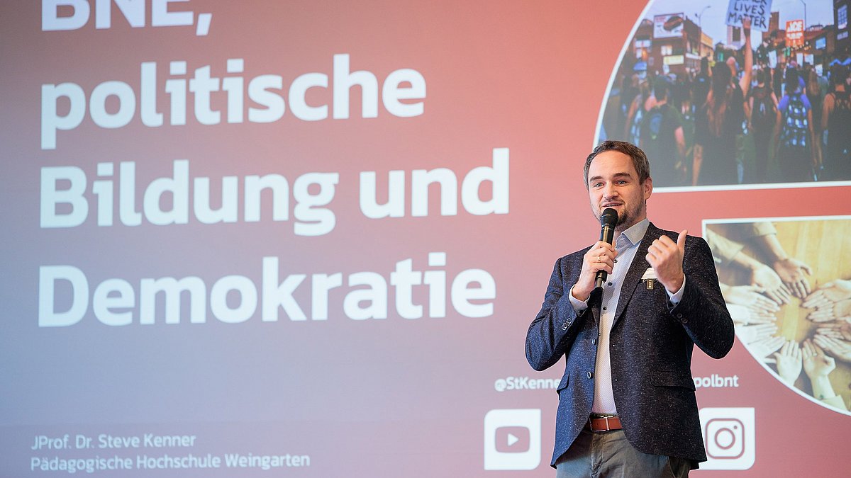 Das Farbfoto zeigt Juniorprofessor Dr. Steve Kenner der Pädagogischen Hochschule Weingarten, der etwas auf einer Bühne präsentiert. Auf seiner Präsentationsfolie steht „BNE, politische Bildung und Demokratie“ und es ist ein Bild einer „Black Lives Matter“-Demonstration sowie eines von Händen, die einen Kreis bilden, zu sehen.